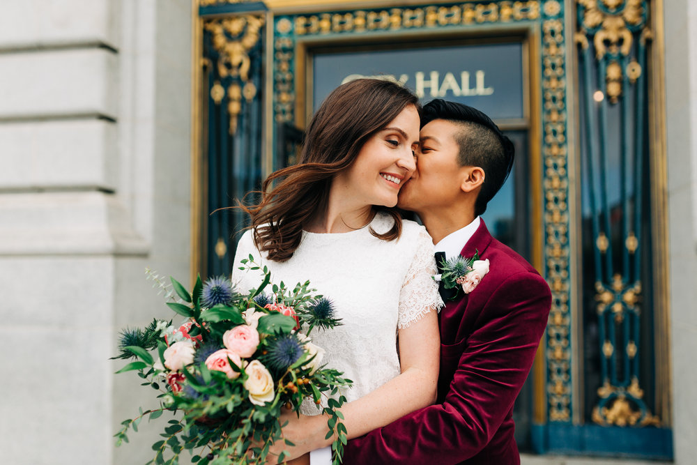 Brides in short white lace dress and burgundy velvet blazer sharing a kiss on San Francisco City Hall steps