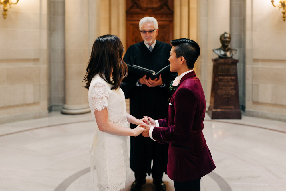 brides in short white dress and burgundy velvet blazer hold hands during their civil ceremony