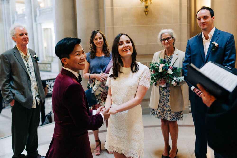 brides in short white dress and burgundy velvet blazer hold hands and family members look over to officiant