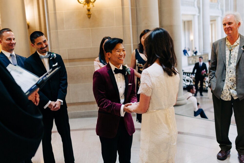 brides in short white dress and burgundy velvet blazer hold hands during their civil ceremony