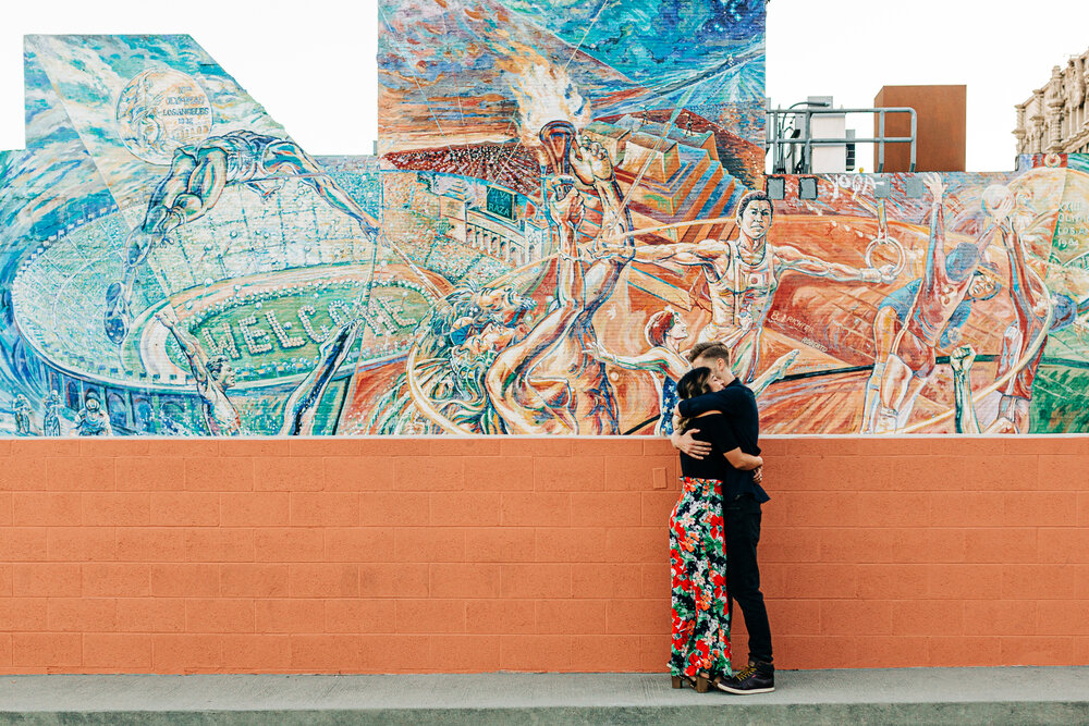 Two engaged people sharing a hug in front of a brightly colored mural in downtown