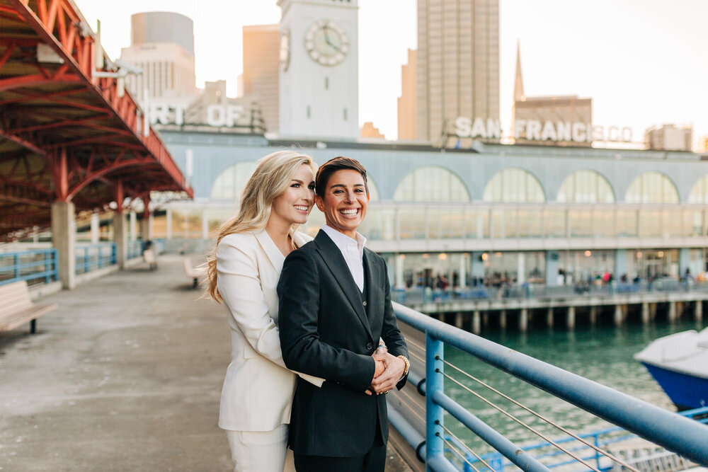  Two brides in tuxes together in front of the San Francisco Ferry Building by the Bay 