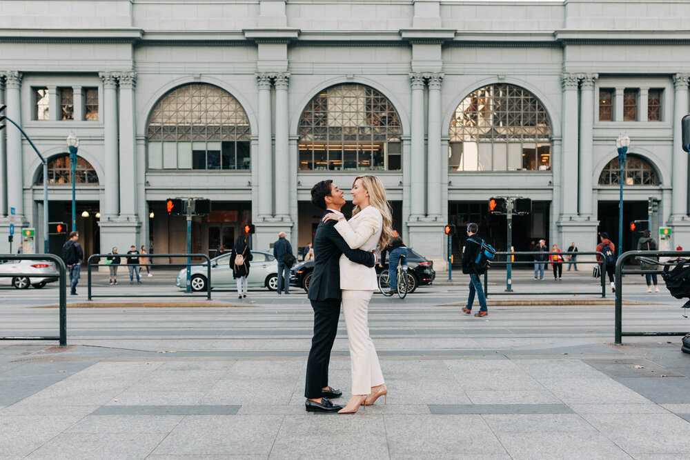 Couple in tuxedos embracing in front of the San Francisco Ferry Building at their Embarcadero elopement 