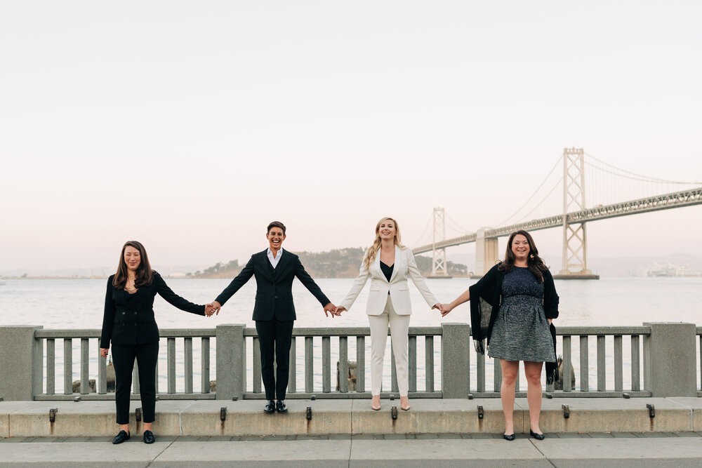  Couple and friends hold hands in front of the Bay Bridge at their Embarcadero elopement 