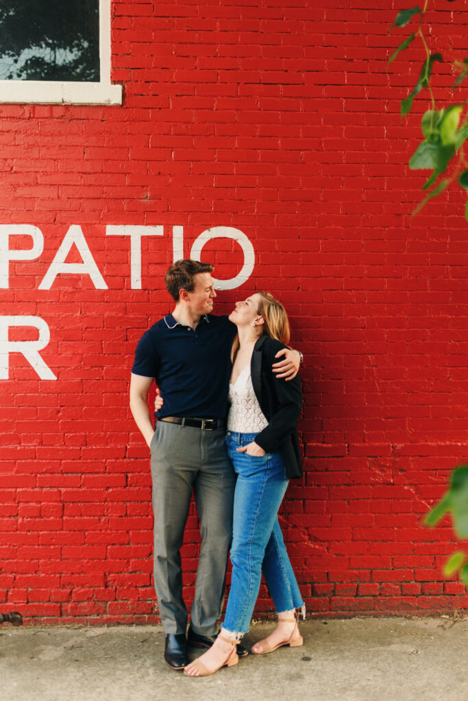 An engaged couple smiling at each other in front of a bright red brick wallalong South Main Street in Memphis