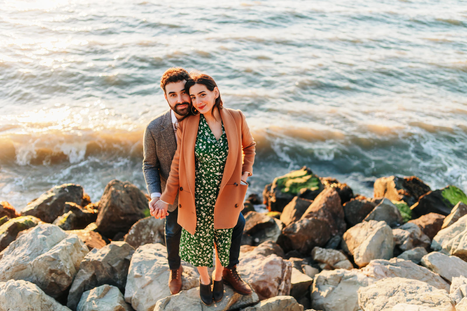 candid photo of a couple standing along rocks by the waterfront and taking in sunset during golden hour
