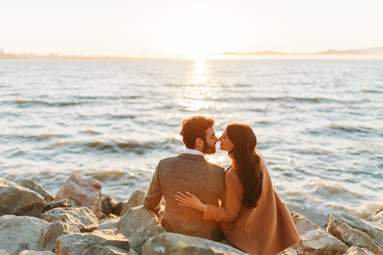 candid photo of a couple sitting along rocks by the waterfront and takes in sunset