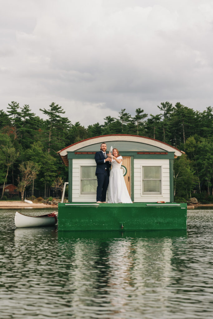 Newlyweds pop champagne off of a houseboat at their New England summer camp wedding